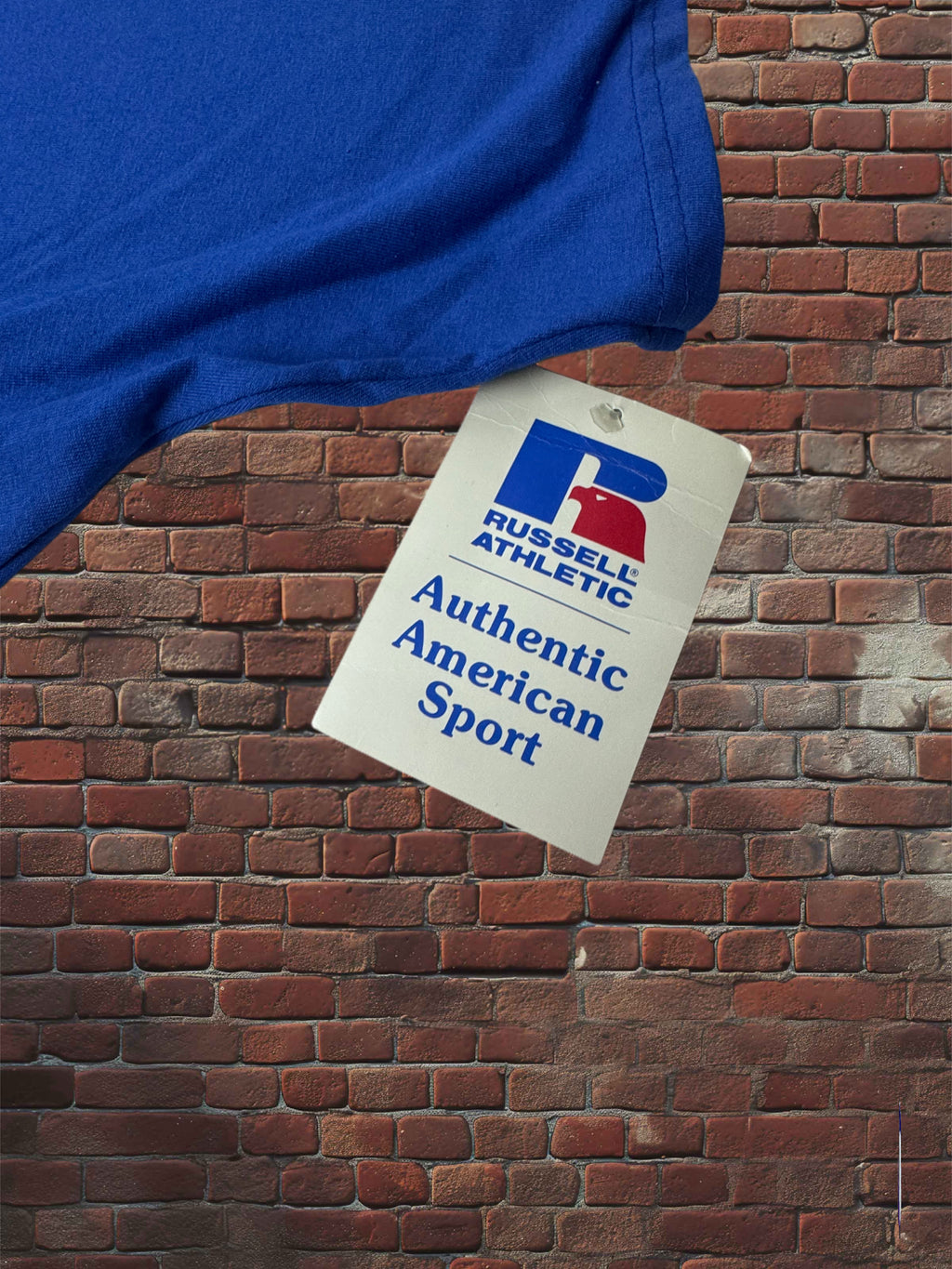 A blue athletic t-shirt is shown draped over a red brick wall, with a white hangtag attached. The tag features the Russell Athletic logo and the text 'Authentic American Sport'.
