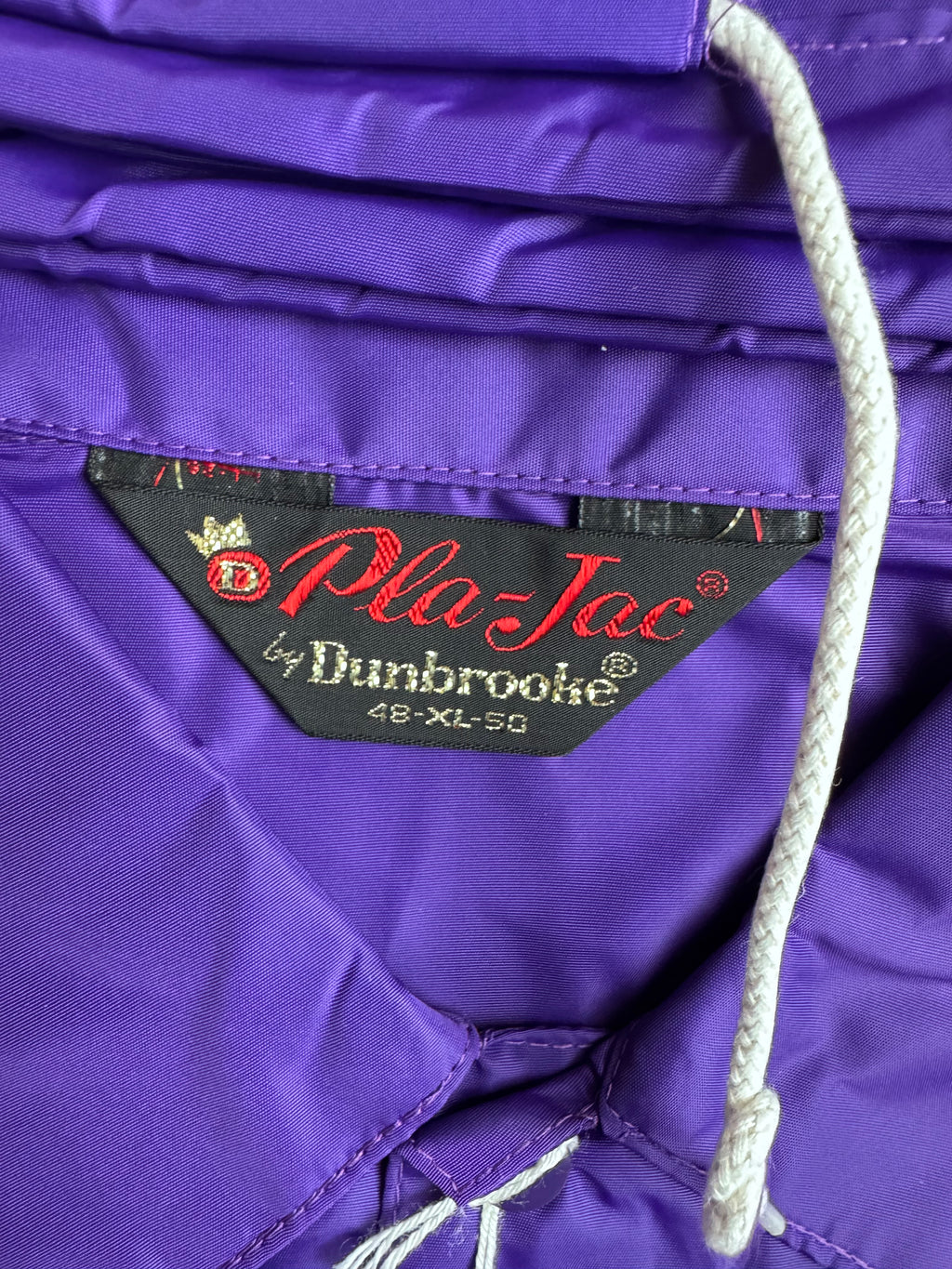 Close-up of a purple nylonjacket showing the interior label which reads 'Pla-Jac by Dunbrooke' and size '48-XL-50', with a white drawstring visible.