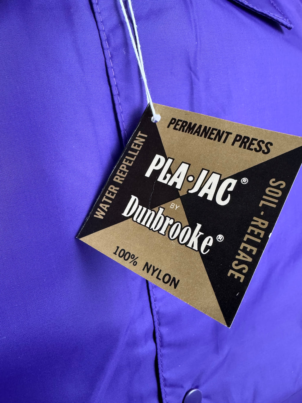 A close-up of a bright purple nylon garment with a hanging PLA-JAC by Dunbrooke permanent press water repellent soil-release tag.
