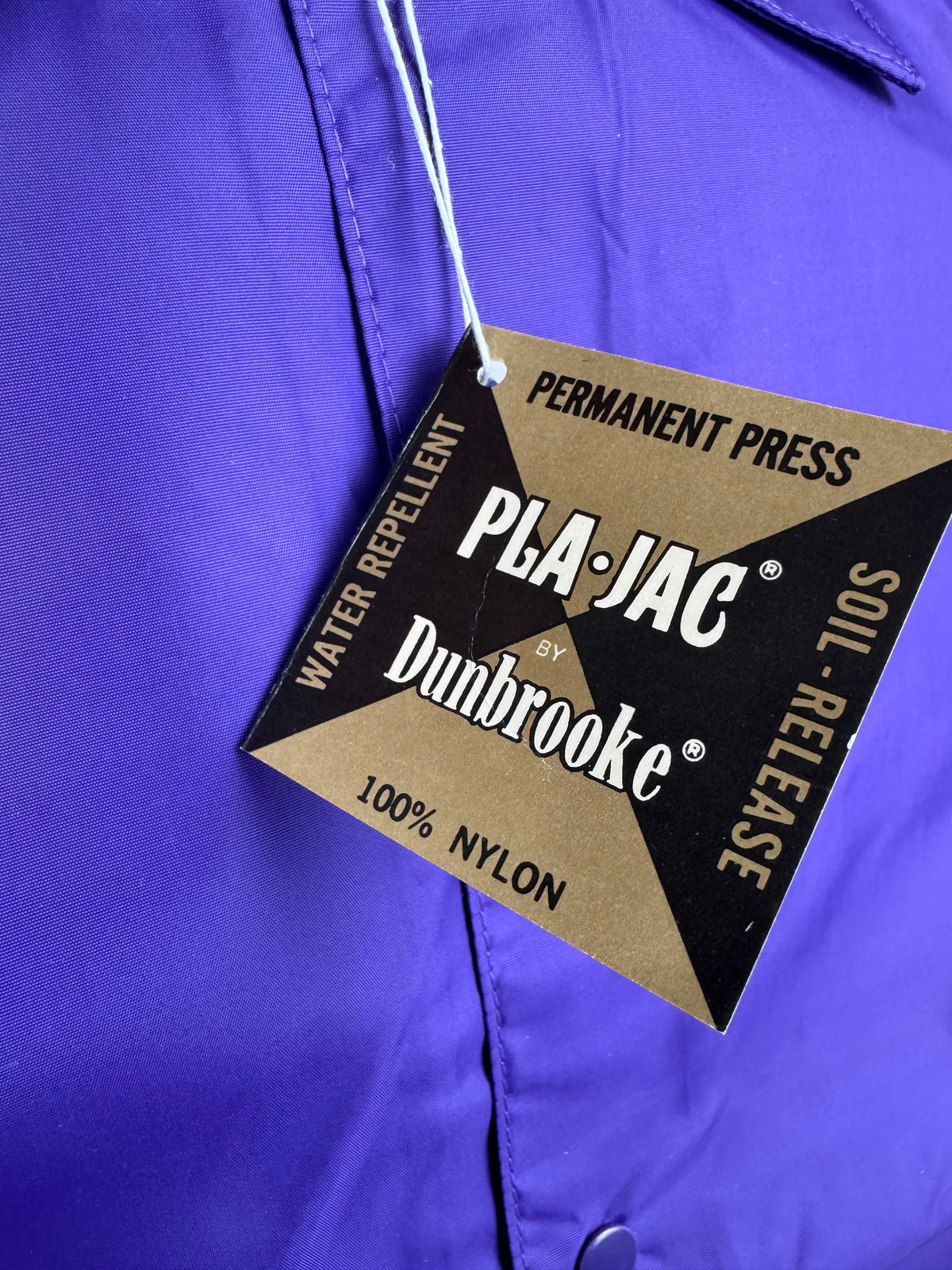 A close-up of a bright purple nylon garment with a hanging PLA-JAC by Dunbrooke permanent press water repellent soil-release tag.