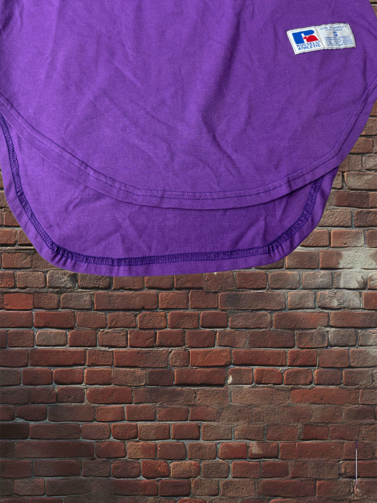 A purple athletic jersey top, cropped to show the curved hem and a small Texas Longhorns logo patch, set against a red-brown brick wall background.