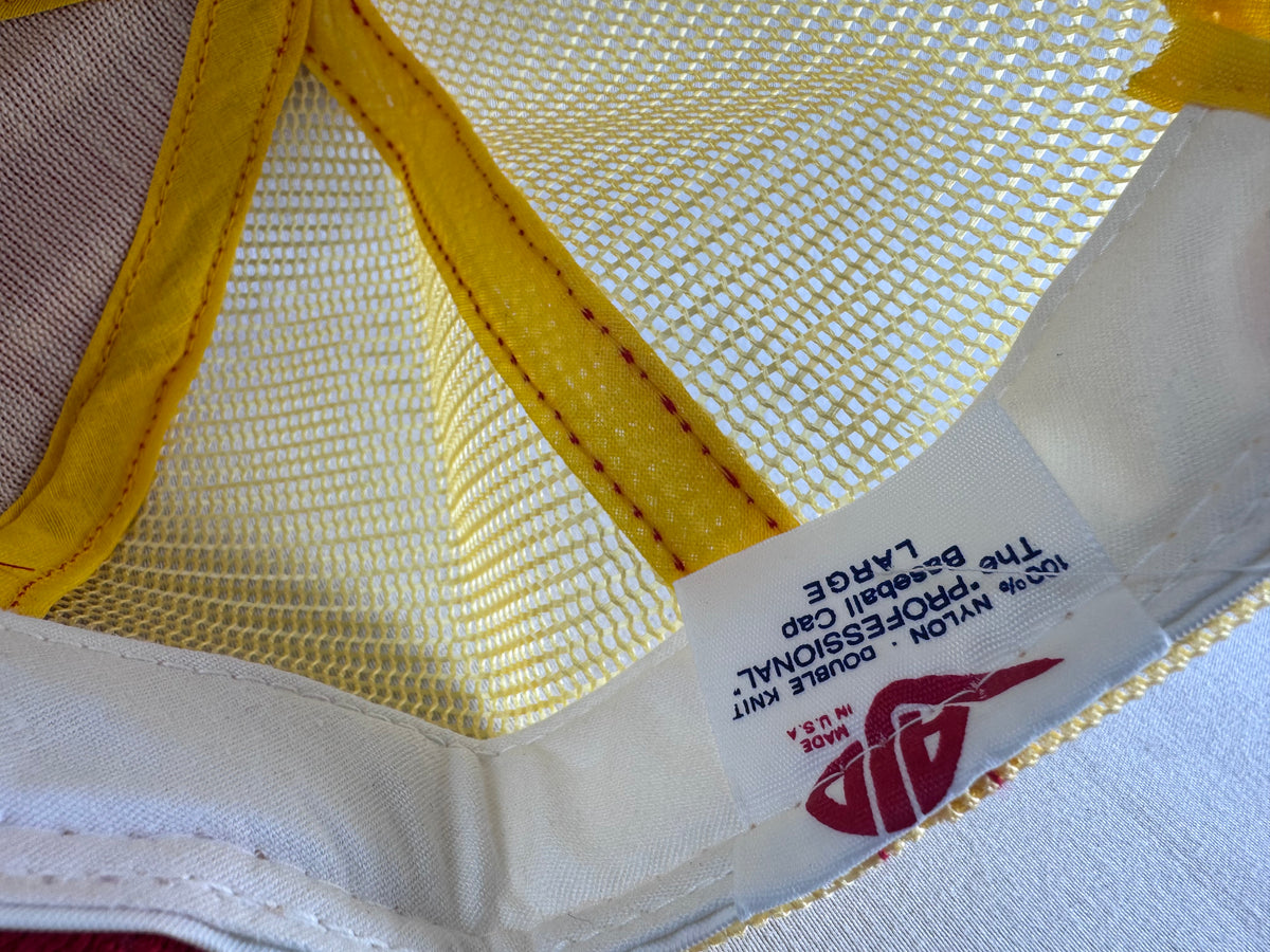 Close-up of the inside of a baseball cap showing a white mesh back panel with yellow trim and a sewn-on size label that reads 100% nylon double knit The Professional Large. AJD brand, Made in the USA