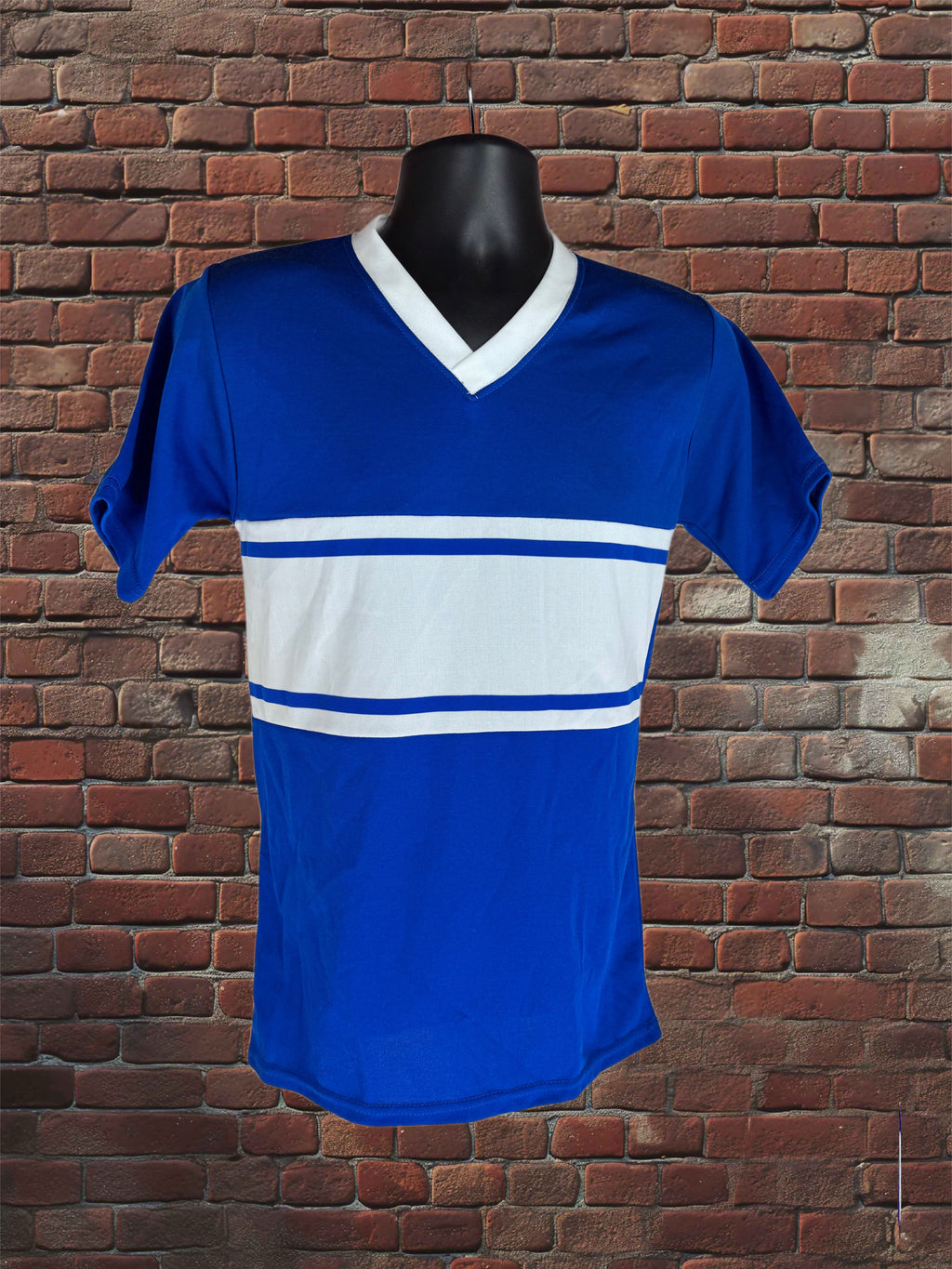 A royal blue short-sleeve V-neck athletic jersey with a wide white horizontal stripe across the chest, displayed on a black torso mannequin against a red-brown brick wall background.