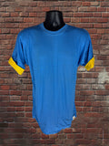 A blue short-sleeve t-shirt with yellow rolled cuffs, displayed on a black mannequin torso against a red brick wall background.