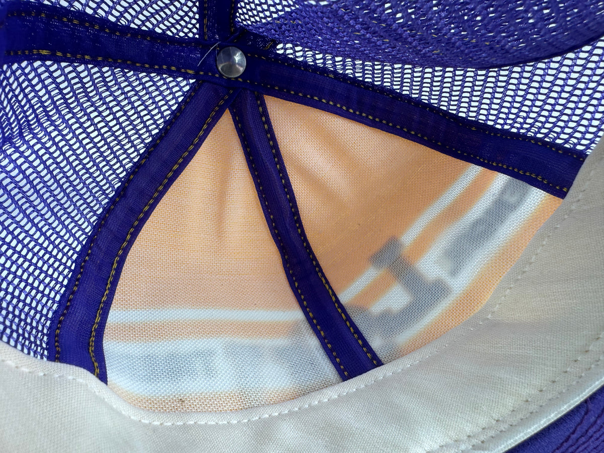 Close-up of the back panels of a trucker-style cap, showing a purple mesh back, purple fabric straps, a silver metal snap, and a white fabric visor edge.