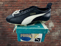 A black and white Puma soccer cleat with white laces, displayed on top of its teal and white Puma shoebox against a red brick wall background.