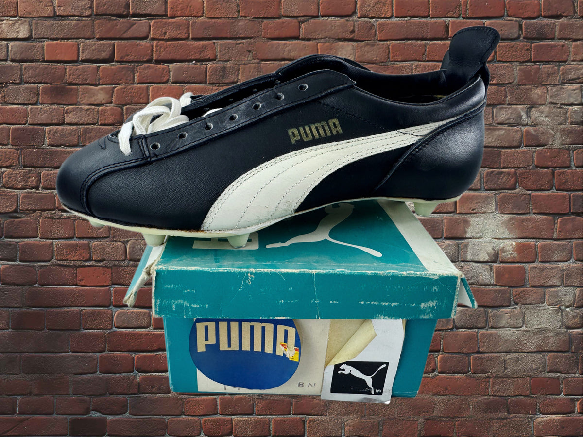 A black and white Puma soccer cleat with white laces, displayed on top of its teal and white Puma shoebox against a red brick wall background.