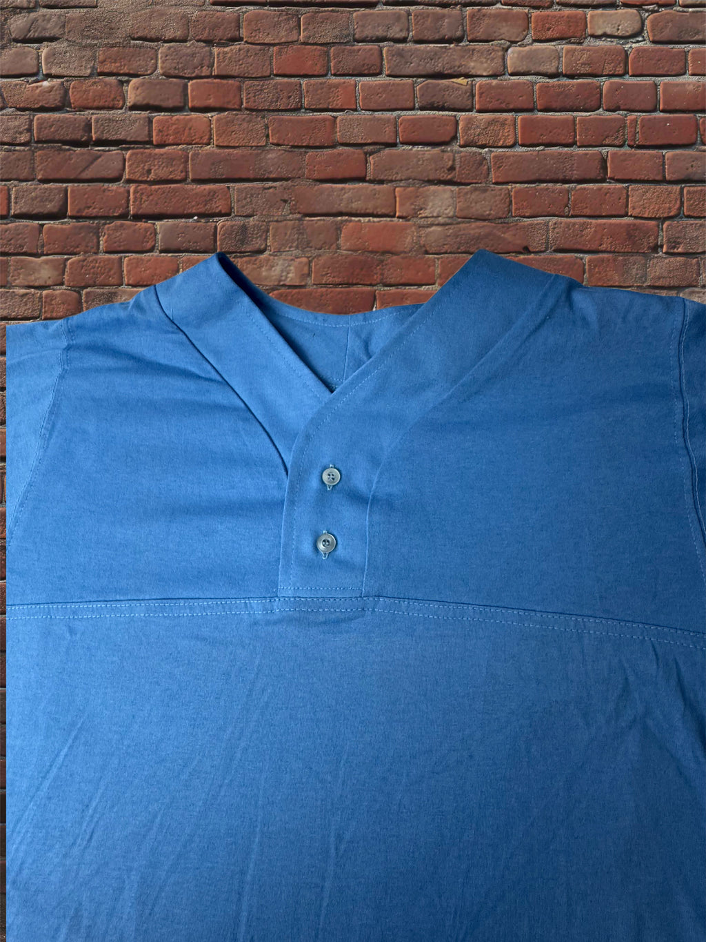 A blue short-sleeve henley shirt with a split collar and two buttons, laid flat against a red brick wall background.