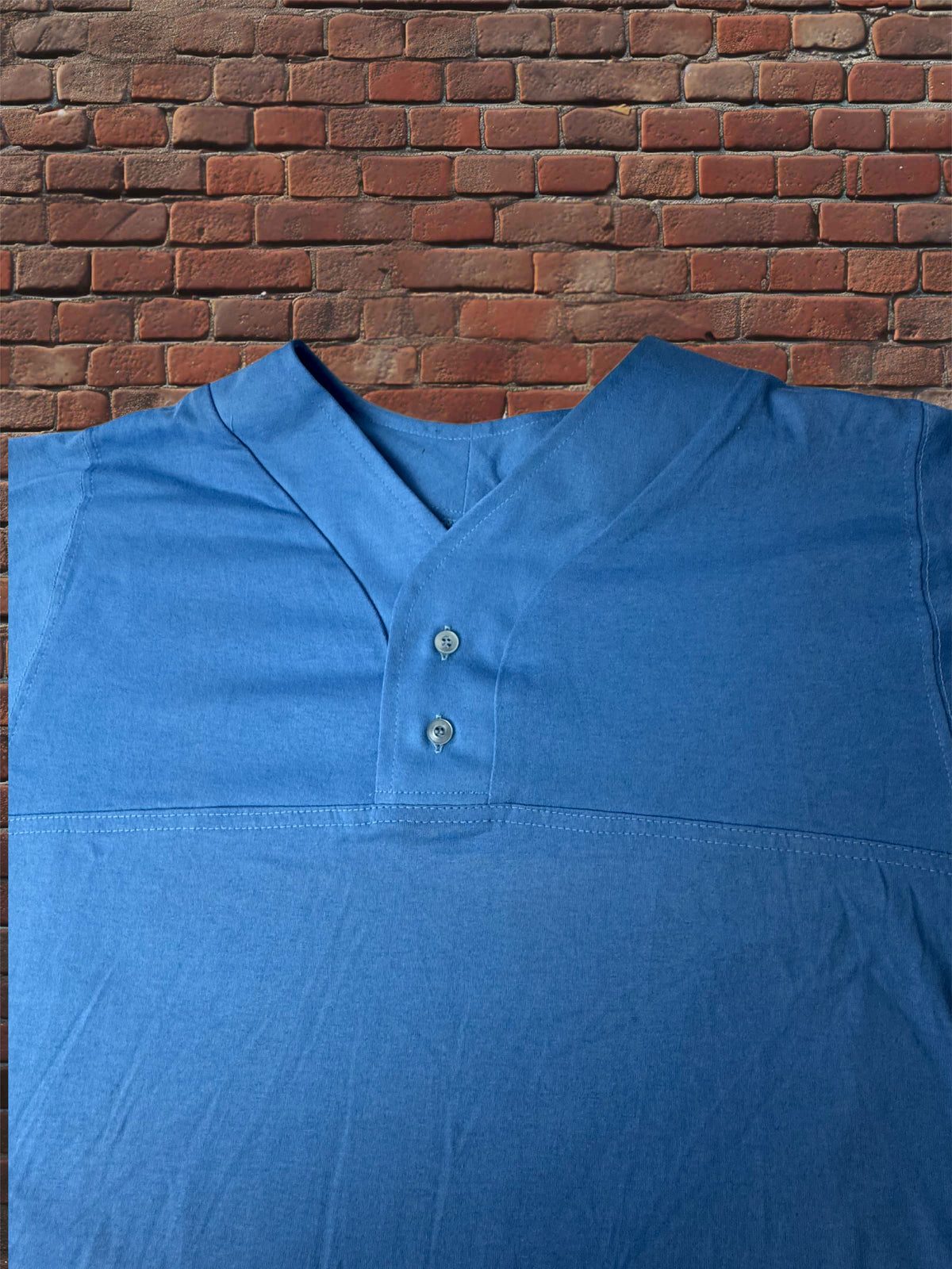 A blue short-sleeve henley shirt with a split collar and two buttons, laid flat against a red brick wall background.
