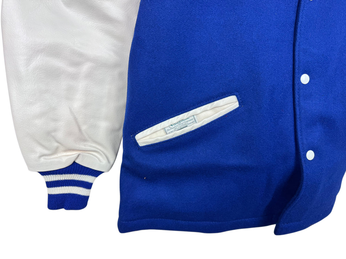 Close-up of a varsity-style jacket showing a royal blue wool body with white snap buttons, a white welt pocket, and a white naugalite sleeve with blue striped ribbed cuffs.