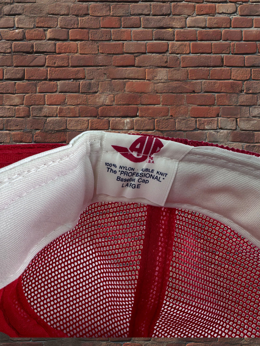 Close-up of the inside crown of a red and white baseball cap, showing the white fabric band, red mesh panel, and a sewn-in label with a stylized 'AJD' logo and text reading 'The Professional Baseball Cap Large'.