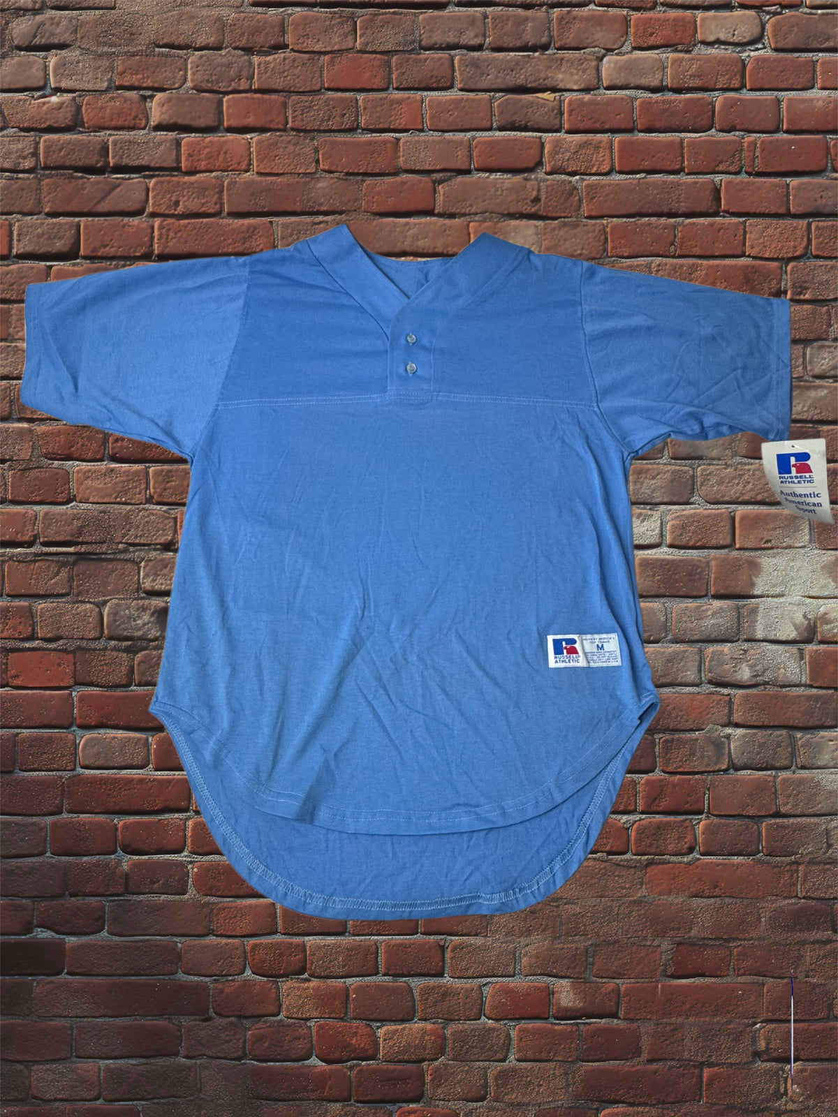 A light blue short-sleeve athletic jersey with a split collar and two-button placket, laid flat against a red-brown brick wall. A small white tag is visible on the right side.