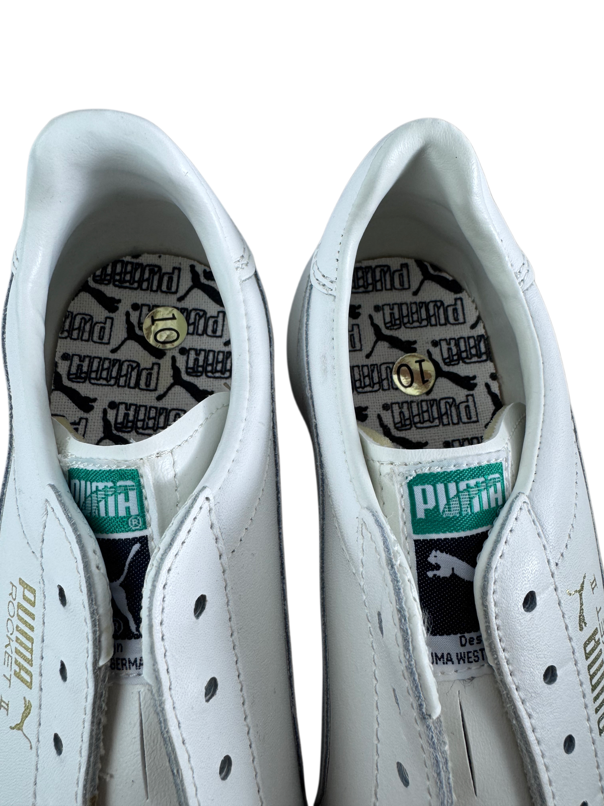 A pair of white Puma soccer/football shoes shown from the side and back, featuring the iconic green Puma cat logo on the heel tab and a black-and-white patterned insole with gold Puma logos.