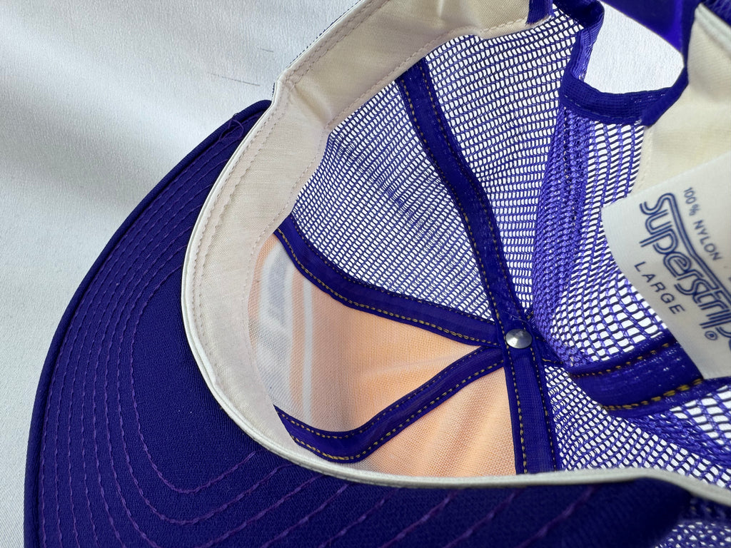 Close-up of the interior of a trucker-style cap, showing a purple visor, purple mesh back panels, a white fabric lining, and a white size/brand tag reading '100% nylon', 'Superstripe' and 'LARGE'.