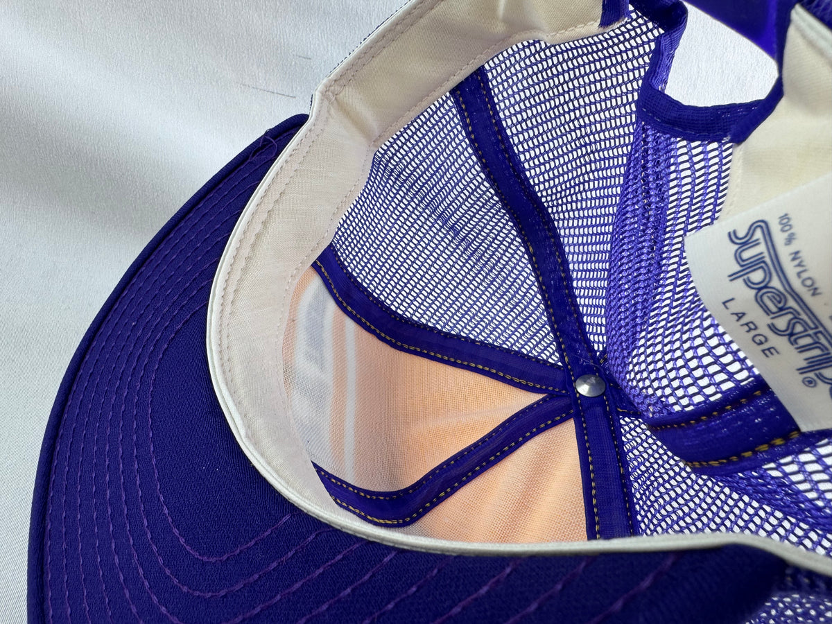 Close-up of the interior of a trucker-style cap, showing a purple visor, purple mesh back panels, a white fabric lining, and a white size/brand tag reading '100% nylon', 'Superstripe' and 'LARGE'.