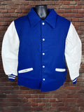 A royal blue and white varsity jacket displayed on a black mannequin torso against a red-brown brick wall background.