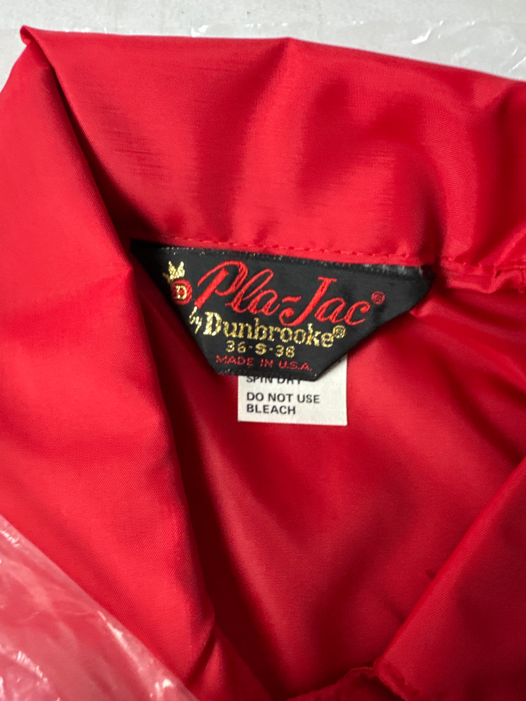 Close-up of a red nylon jacket interior showing a black fabric brand label that reads 'Pla-Jac by Dunbrooke 36 38 Made in USA' with a white 'Spin Dry Do Not Use Bleach' tag below it.