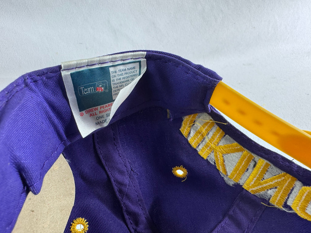 Close-up of a purple baseball cap with a yellow brim, featuring a visible Team NFL brand label on the inside and gold embroidered lettering on the side.