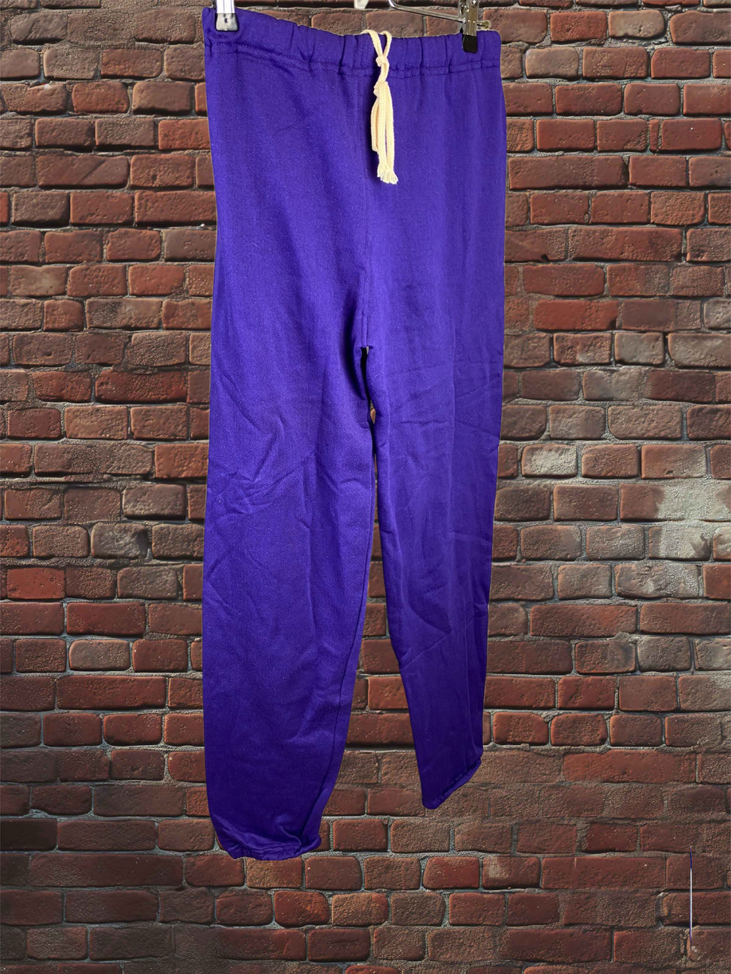 Purple sweatpants with a cream drawstring, hanging against a red brick wall background.