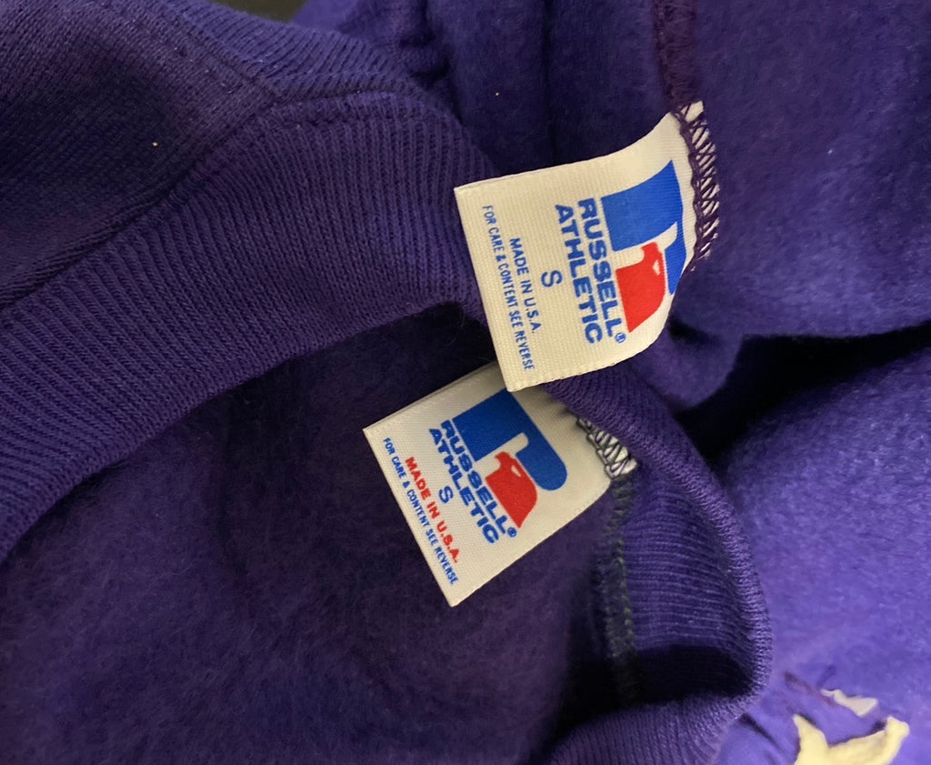 Close-up of a purple knit garment showing two white Russell Athletic interior tags with the brand logo in blue and red.
