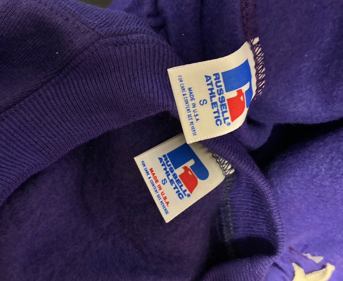 Close-up of a purple knit garment showing two white Russell Athletic interior tags with the brand logo in blue and red.