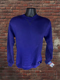 A purple long-sleeve crewneck sweatshirt displayed on a black mannequin torso against a red-brown brick wall background. A hangtag is visible on the right sleeve cuff.