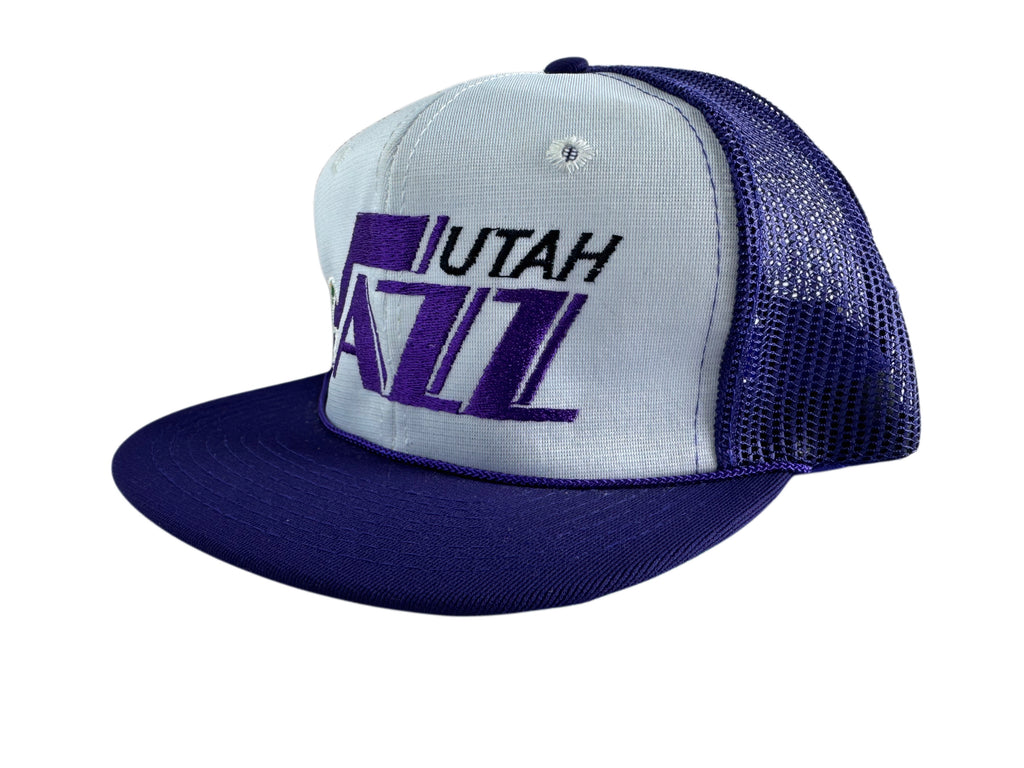 A two-tone Utah Jazz trucker hat featuring a white front panel with purple and black embroidered logo, a dark purple flat brim, and a dark purple mesh back.