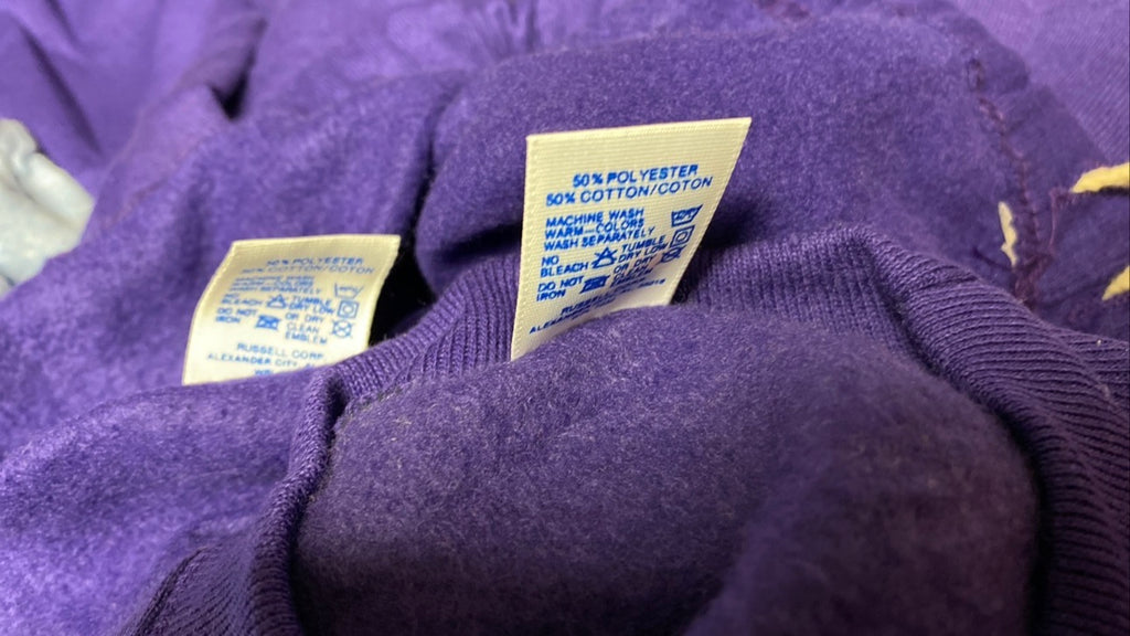 Close-up of a purple knit garment showing two cream-colored care labels with blue text, including one that reads '50% POLYESTER 50% COTTON/CLOTH'.