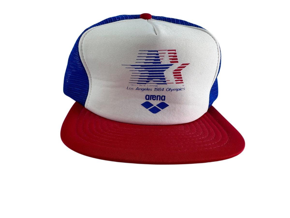 A red, white, and blue trucker hat featuring the Los Angeles 1984 Olympics logo on the front panel and the Arena brand logo below it, set against a plain white background.
