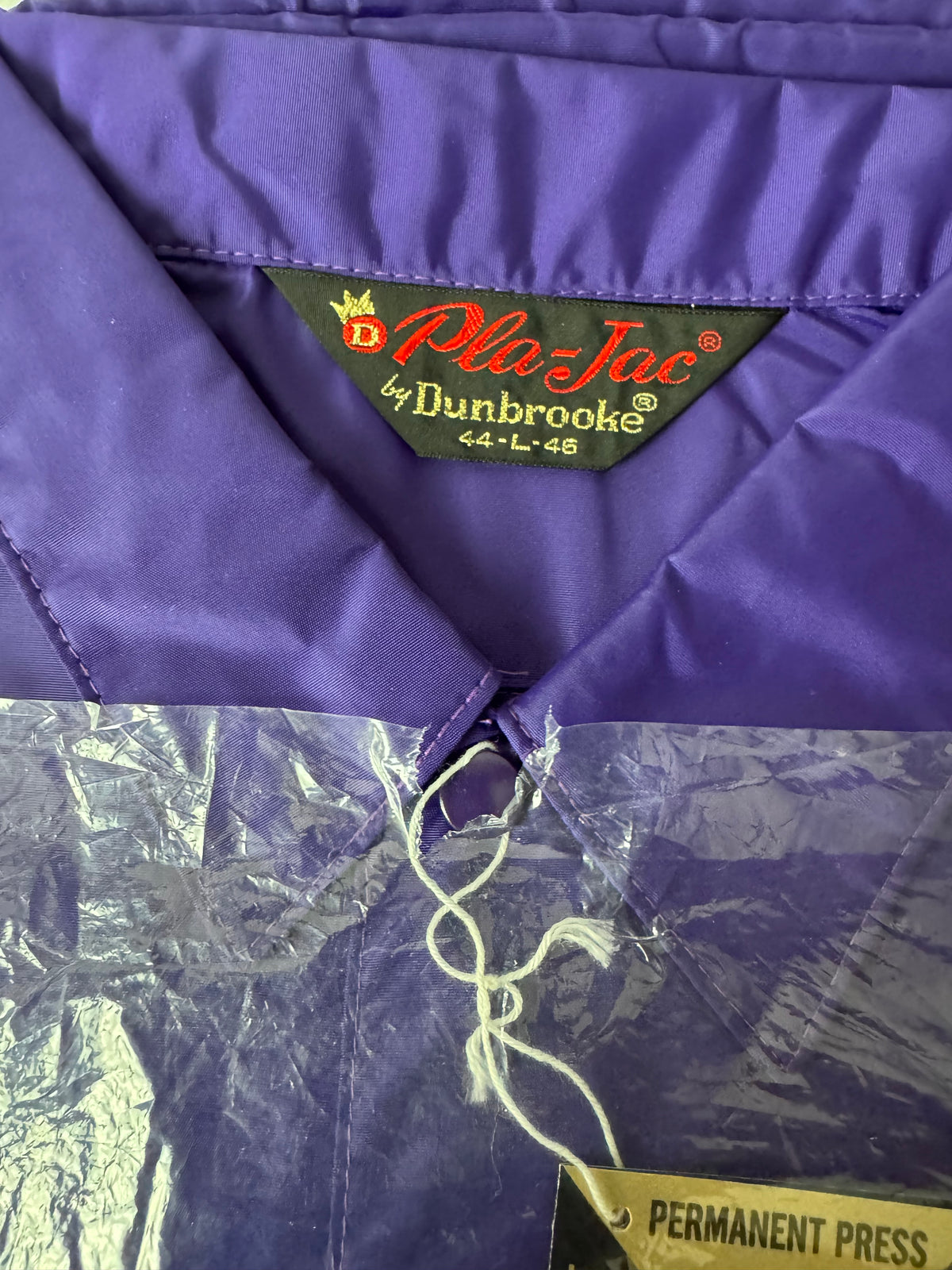 Close-up of a purple nylon jacket collar showing the green Dunbrooke Pla-Jac brand label and a crinkled plastic protective cover.