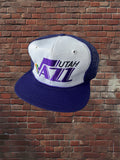 A Utah Jazz trucker hat with a white front panel, purple mesh back, and purple flat brim, featuring the Utah Jazz logo embroidered on the front, set against a red brick wall background.