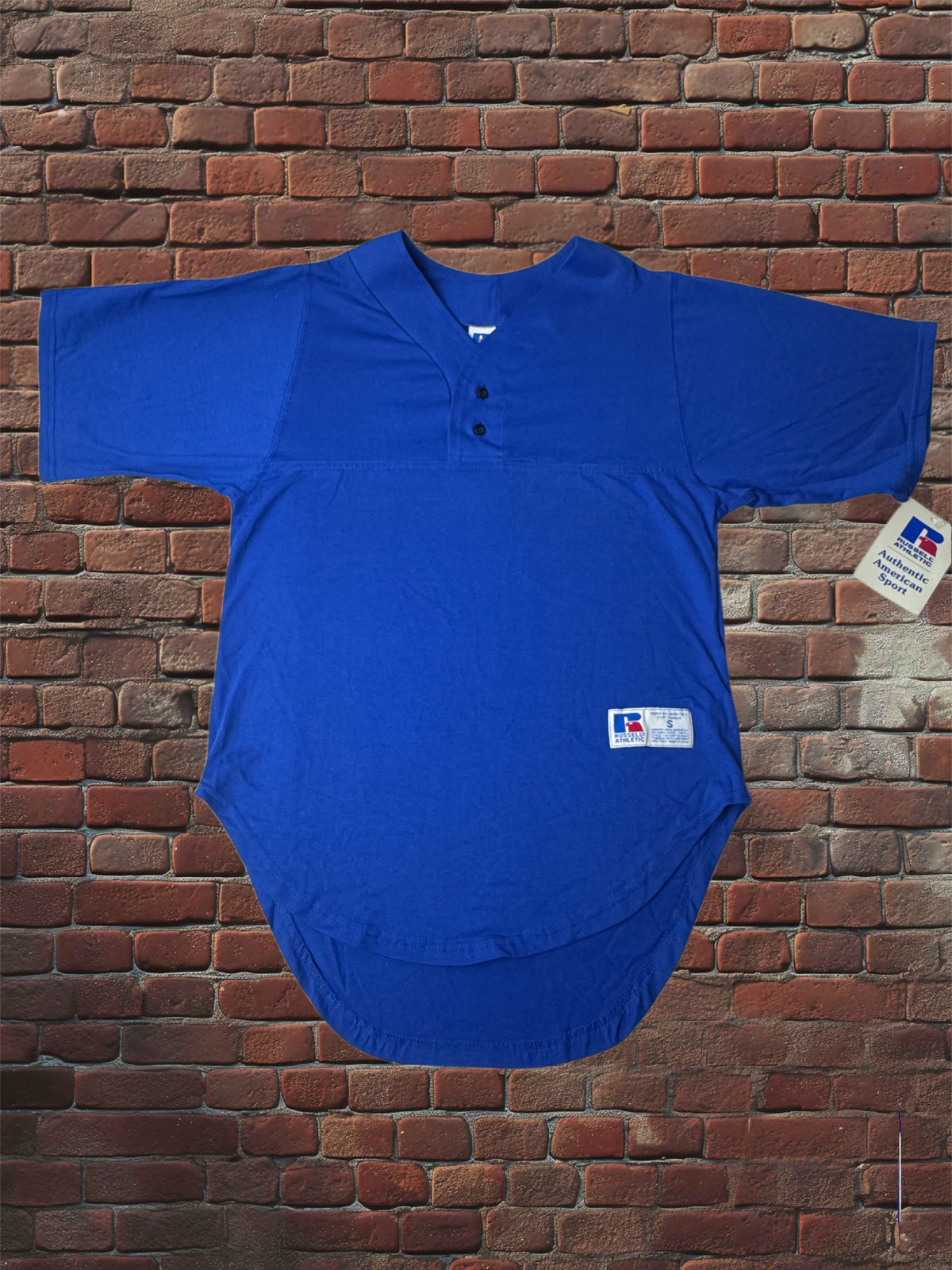 A royal blue short-sleeve baseball jersey top with a three-button henley collar, displayed flat against a red-brown brick wall background. A white Russell Athletic logo patch is visible on the lower front, and a retail tag is attached to the right sleeve.