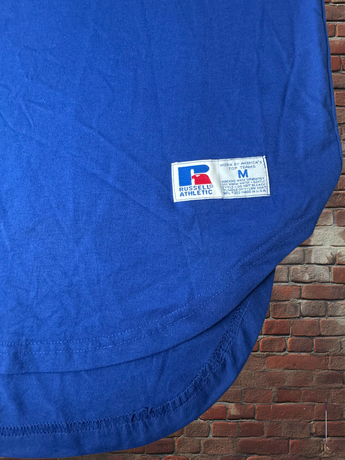 A close-up of a blue athletic t-shirt showing the 'Russell Athletic' brand label, draped over a brick wall background.