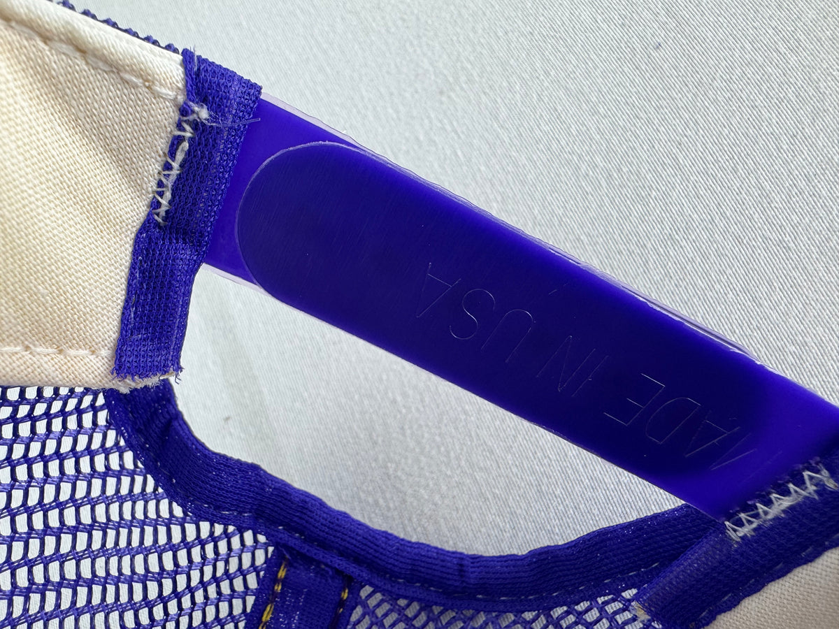 Close-up detail of a trucker hat showing a cream canvas side panel, a purple mesh back, and a translucent royal blue plastic underbrim with 'MADE IN USA' debossed.