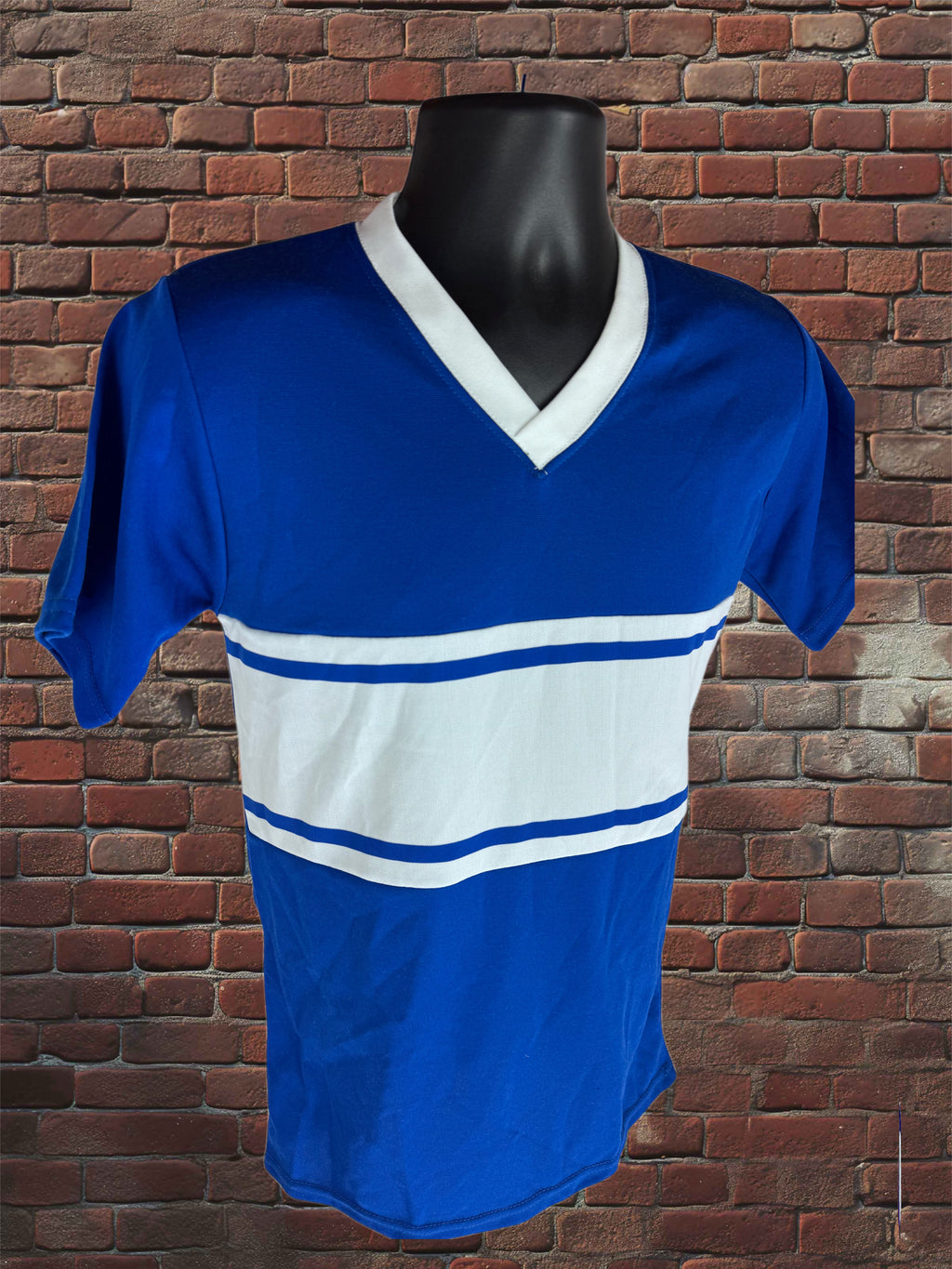 A royal blue short-sleeve V-neck athletic jersey with a wide white chest stripe and thin blue stripes, displayed on a black mannequin torso against a red brick wall background.