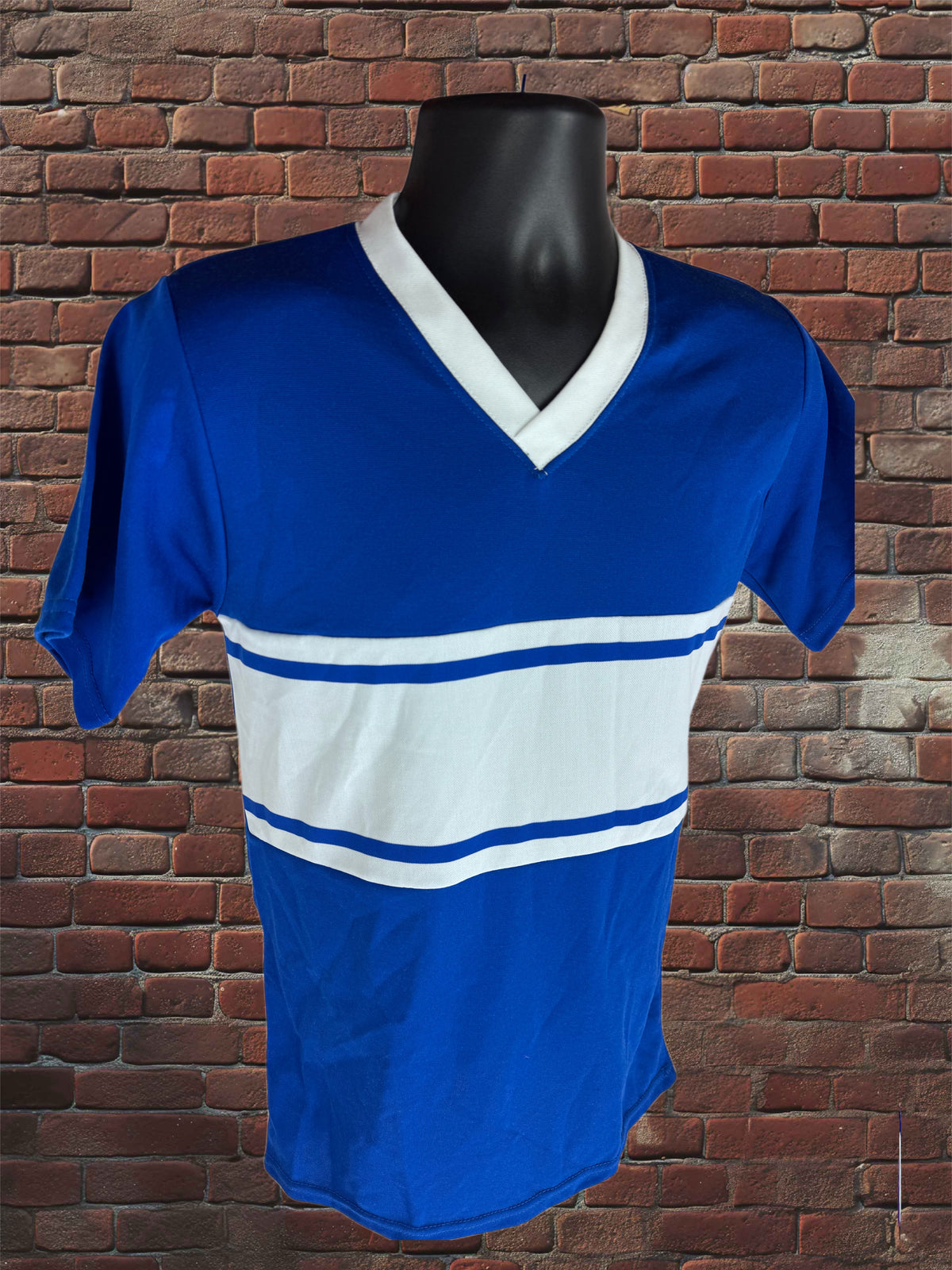 A royal blue short-sleeve V-neck athletic jersey with a wide white chest stripe and thin blue stripes, displayed on a black mannequin torso against a red brick wall background.