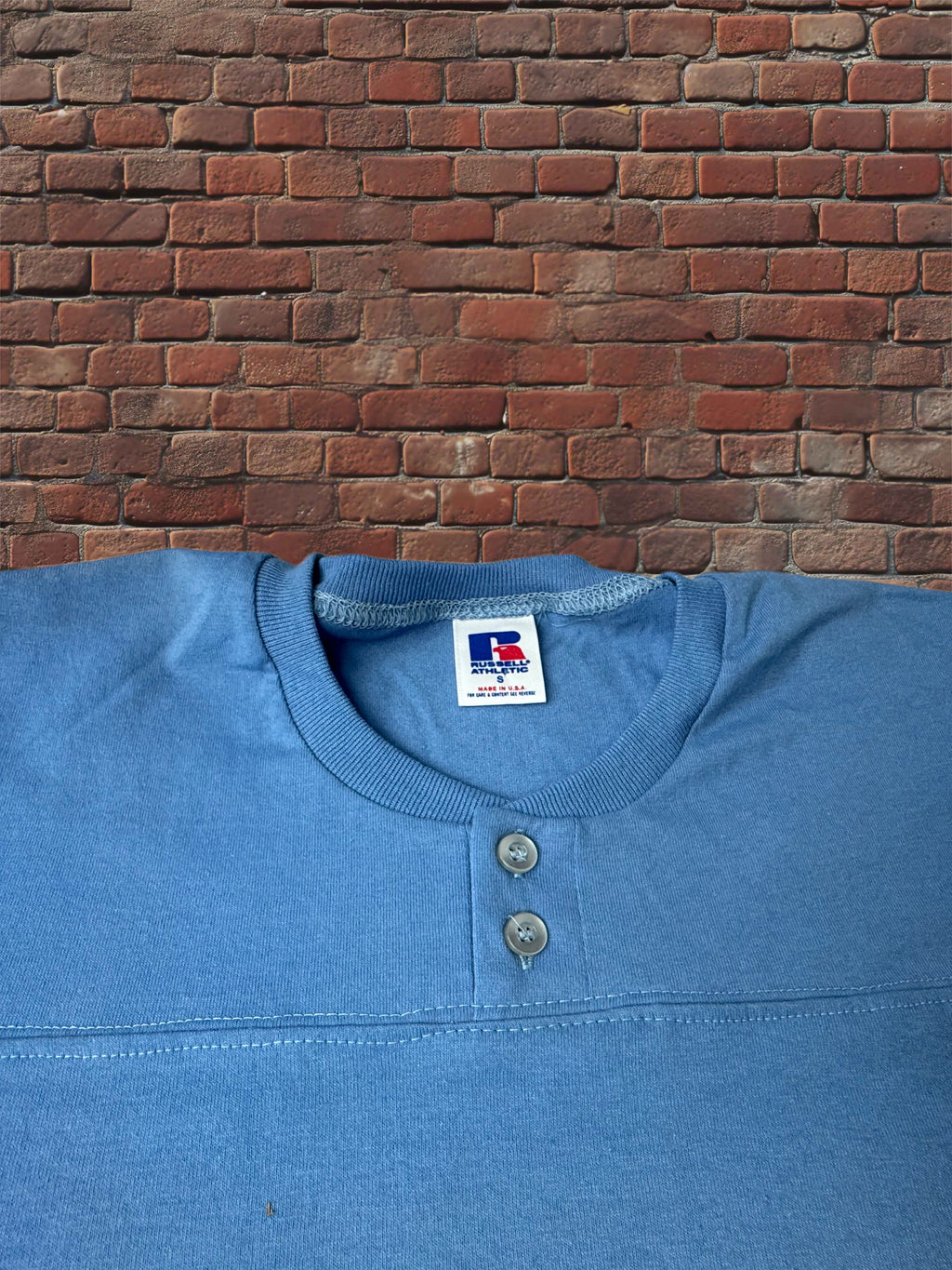 A blue Henley shirt with a two-button placket, laid flat against a red brick wall background. The inner neck label shows the brand 'Russell Athletic'.