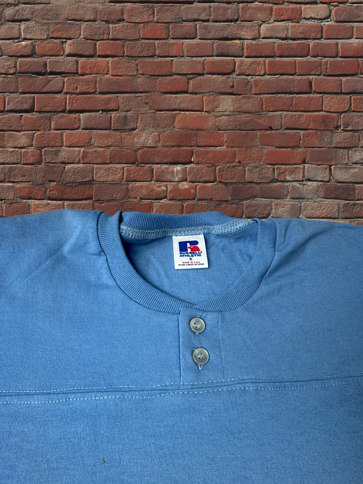 A blue Henley shirt with a two-button placket, laid flat against a red brick wall background. The inner neck label shows the brand 'Russell Athletic'.