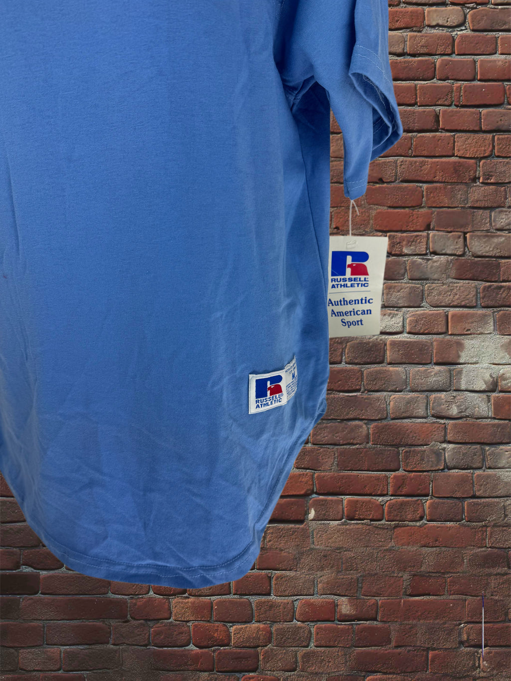 A blue short-sleeve athletic T-shirt hanging against a red brick wall, with a visible Russell Athletic Authentic American Sport hangtag and brand patch.
