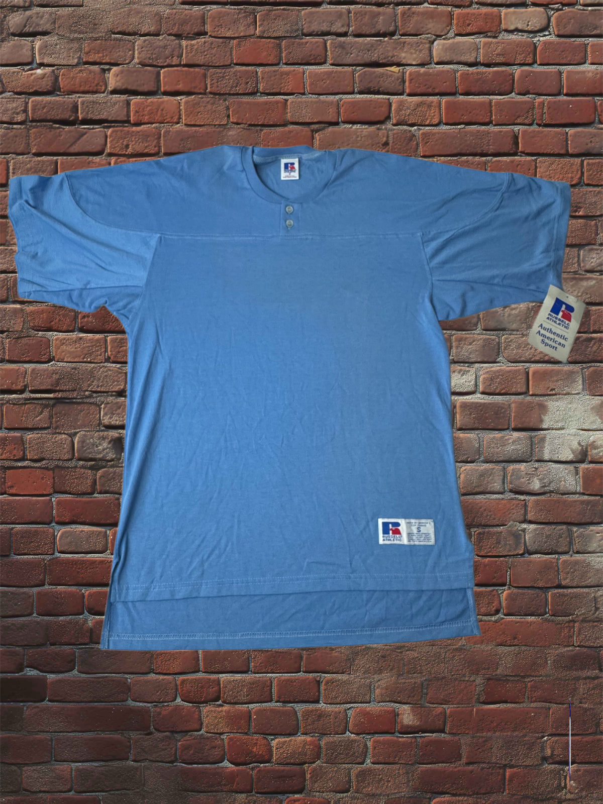 A light blue short-sleeve henley t-shirt laid flat against a red-brown brick wall background. The shirt features a small button placket at the collar, a small rectangular brand tag near the lower hem, and a larger hangtag on the right side.