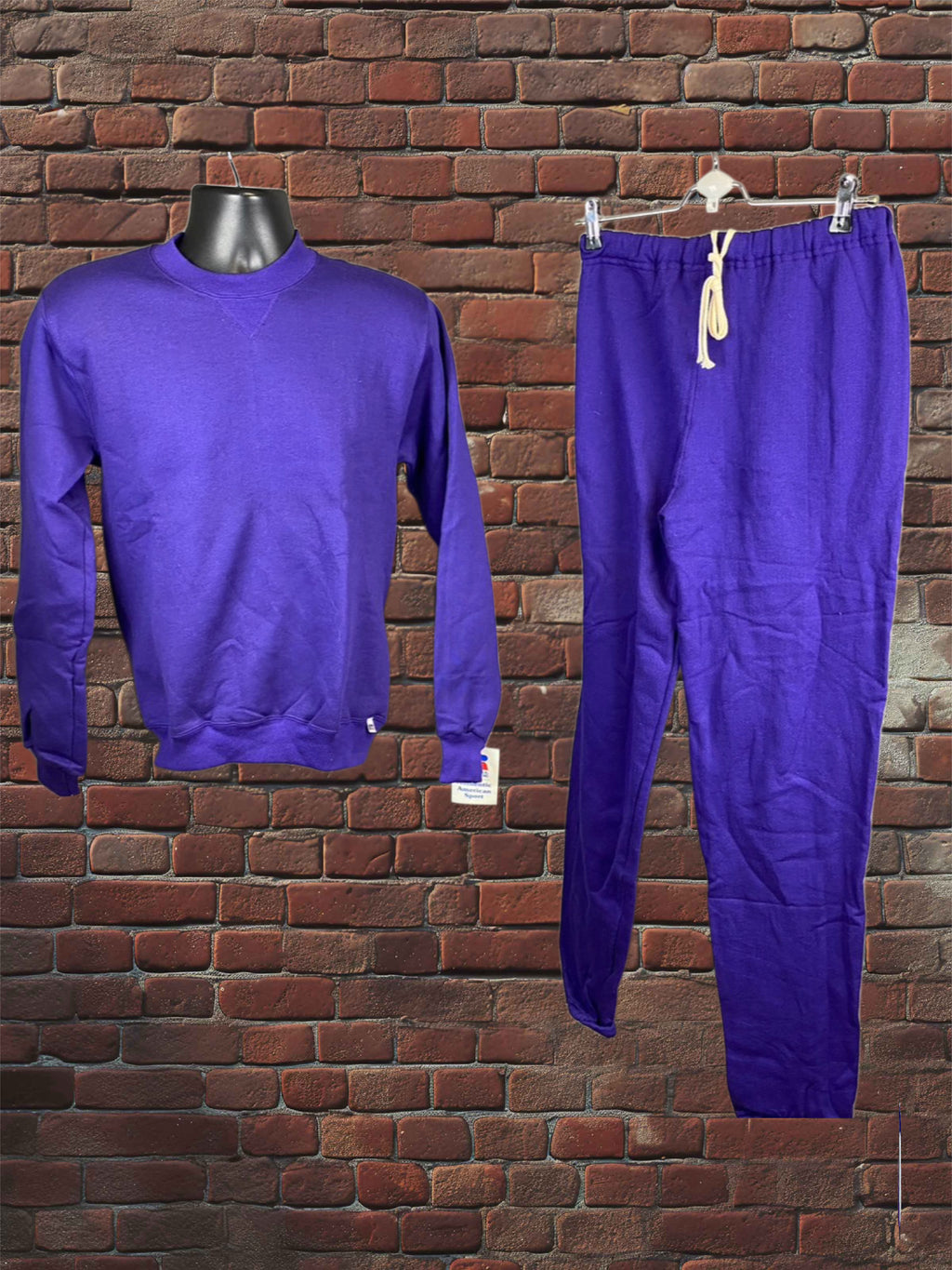 A matching two-piece tracksuit in a vibrant purple color, featuring a long-sleeve crewneck sweatshirt on a black mannequin torso and matching sweatpants hanging from a white hanger, set against a red brick wall background.