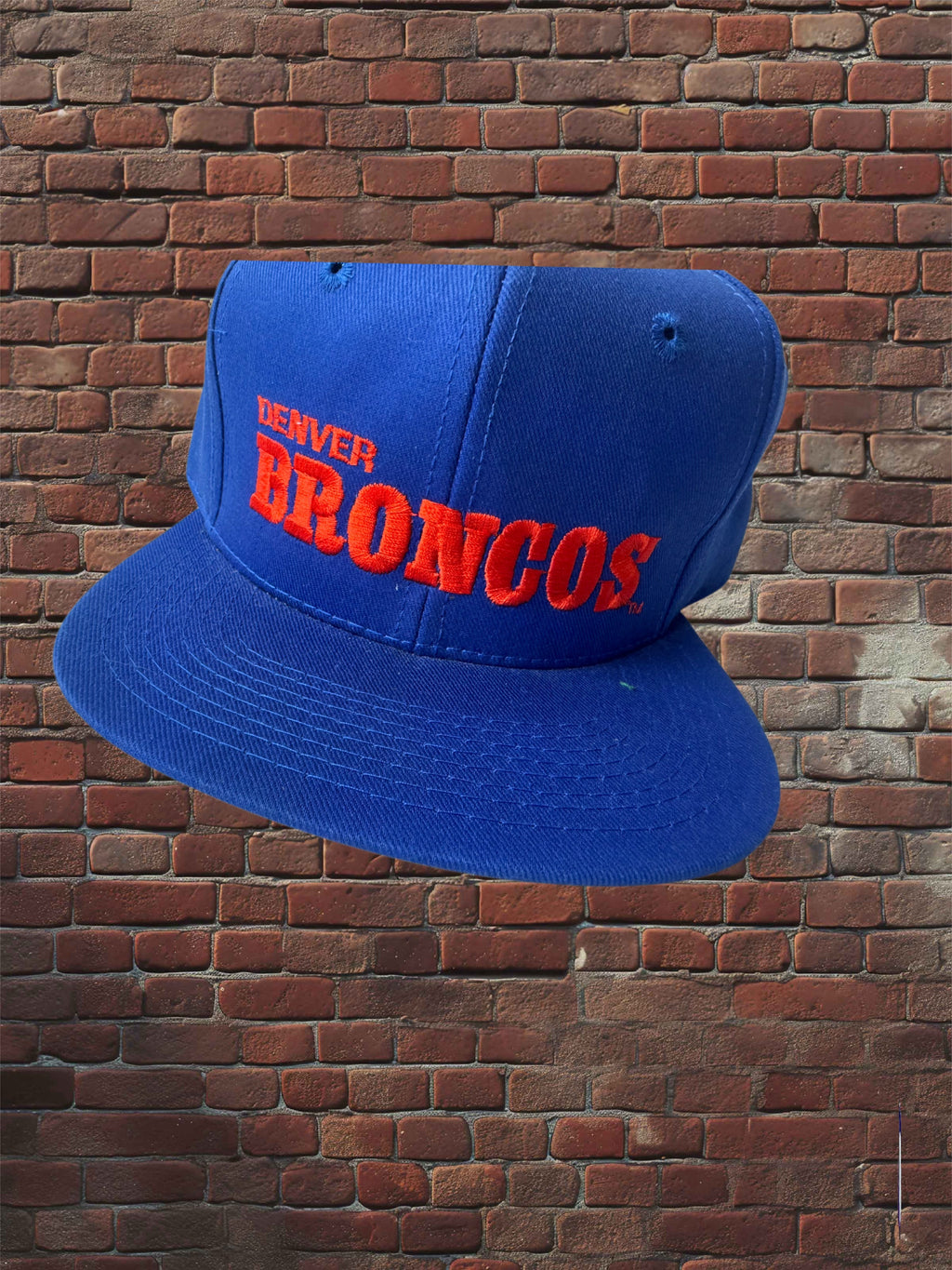 A royal blue snapback cap with 'DENVER BRONCOS' embroidered in red and orange on the front, shown against a red-brown brick wall background.