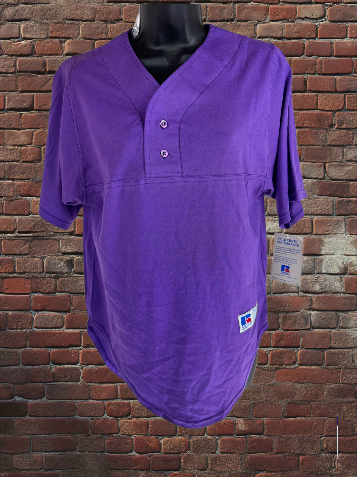A purple short-sleeve baseball-style henley shirt displayed on a black mannequin torso against a red-brown brick wall. The shirt has a split V-neck, two-button placket, and a small white brand tag near the lower right hem.