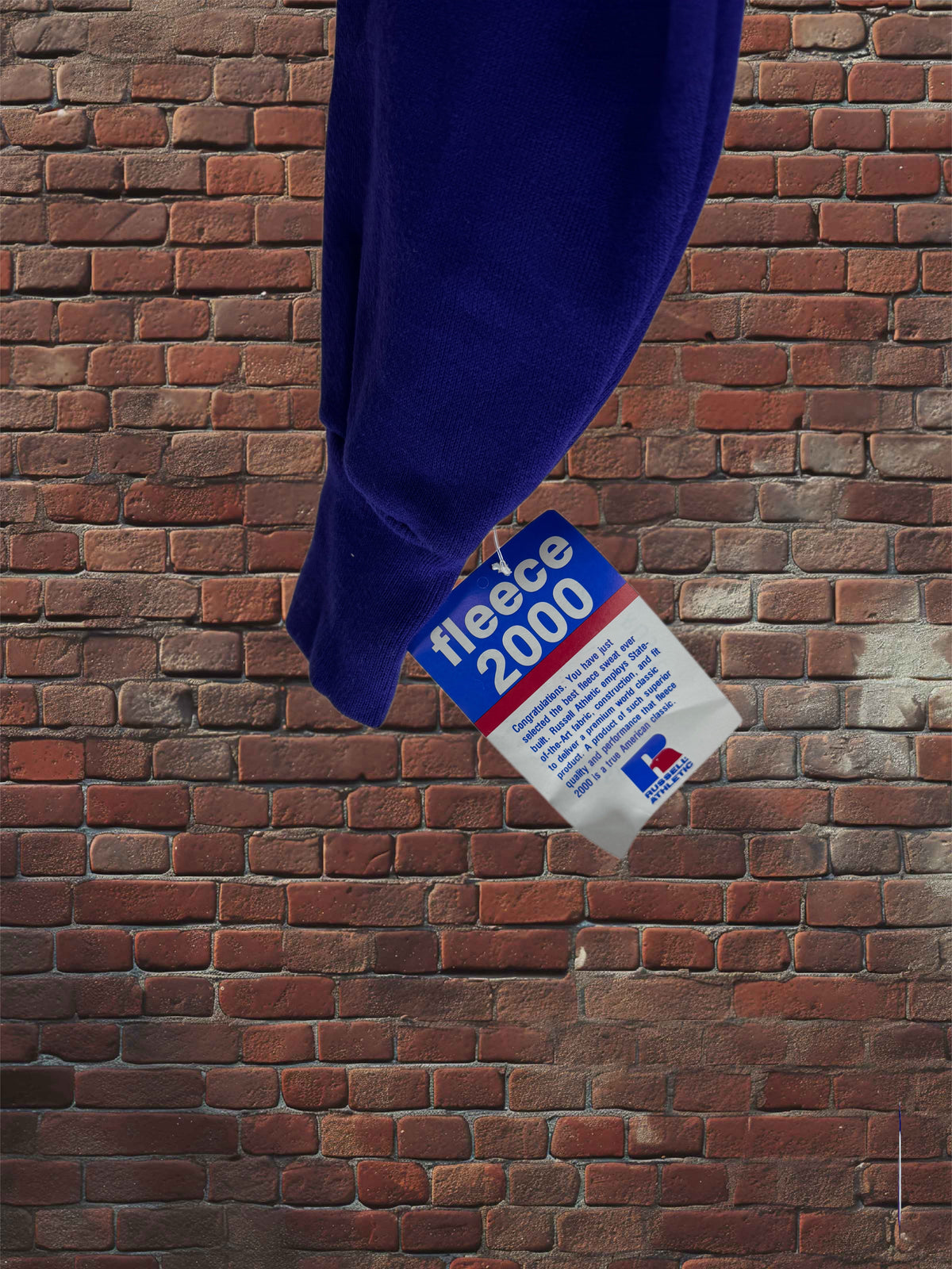 A dark blue fleece garment hangs against a red-brown brick wall, with a hanging fabric tag reading 'fleece 2000' and a small Russell Athletic logo.