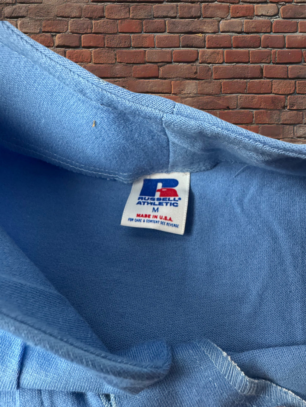Close-up of the inside collar of a light blue Russell Athletic sweatshirt, showing the brand tag and brick wall background.