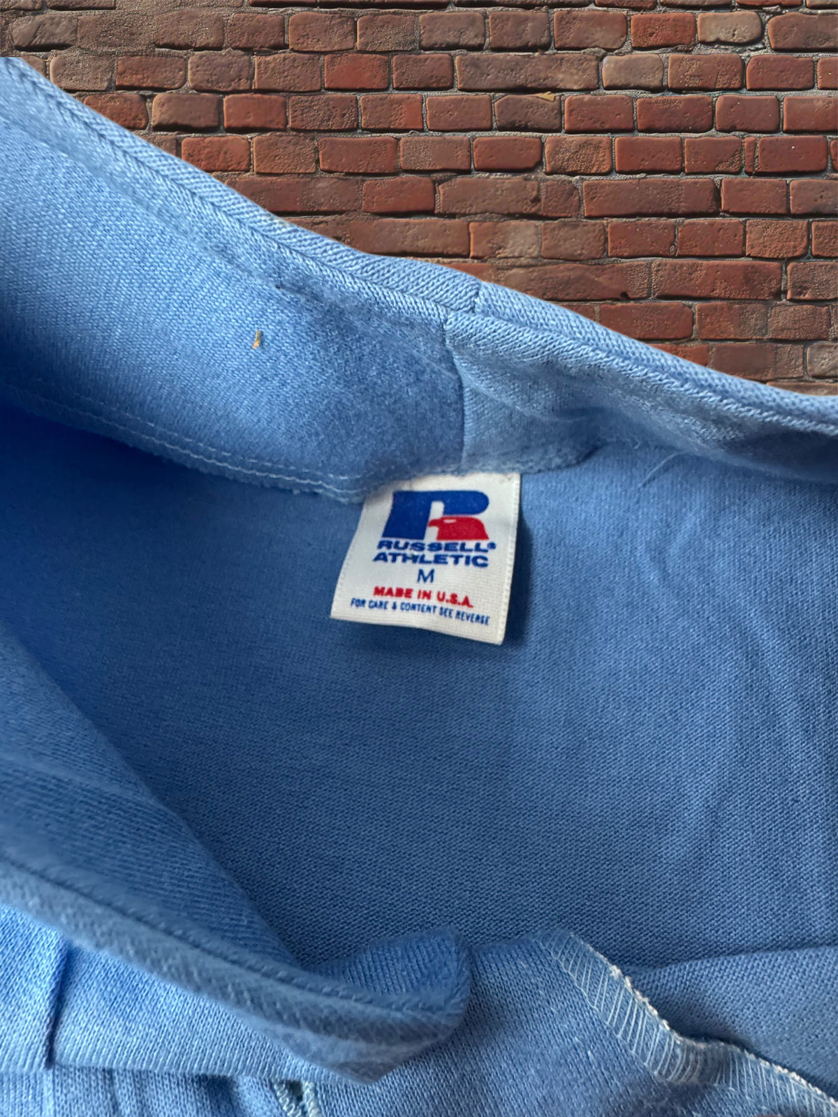 Close-up of the inside collar of a light blue Russell Athletic sweatshirt, showing the brand tag and brick wall background.