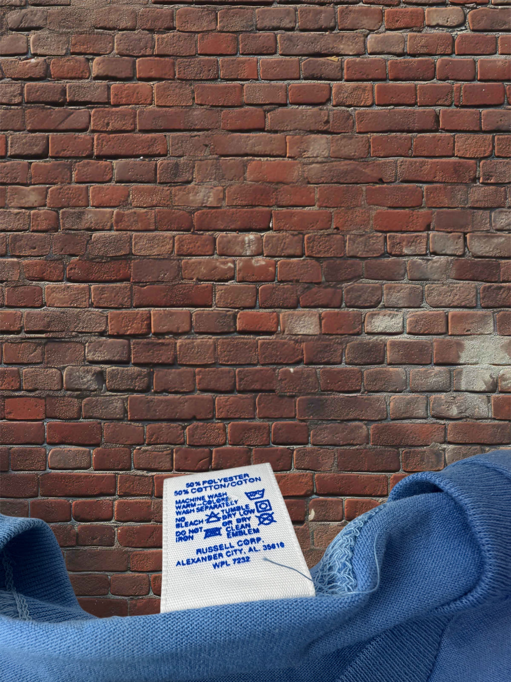 A close-up of a blue sweatshirt showing the inner collar and printed care label, positioned in front of a red-brown brick wall.