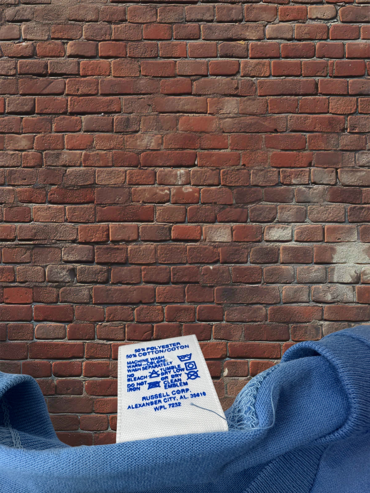 A close-up of a blue sweatshirt showing the inner collar and printed care label, positioned in front of a red-brown brick wall.