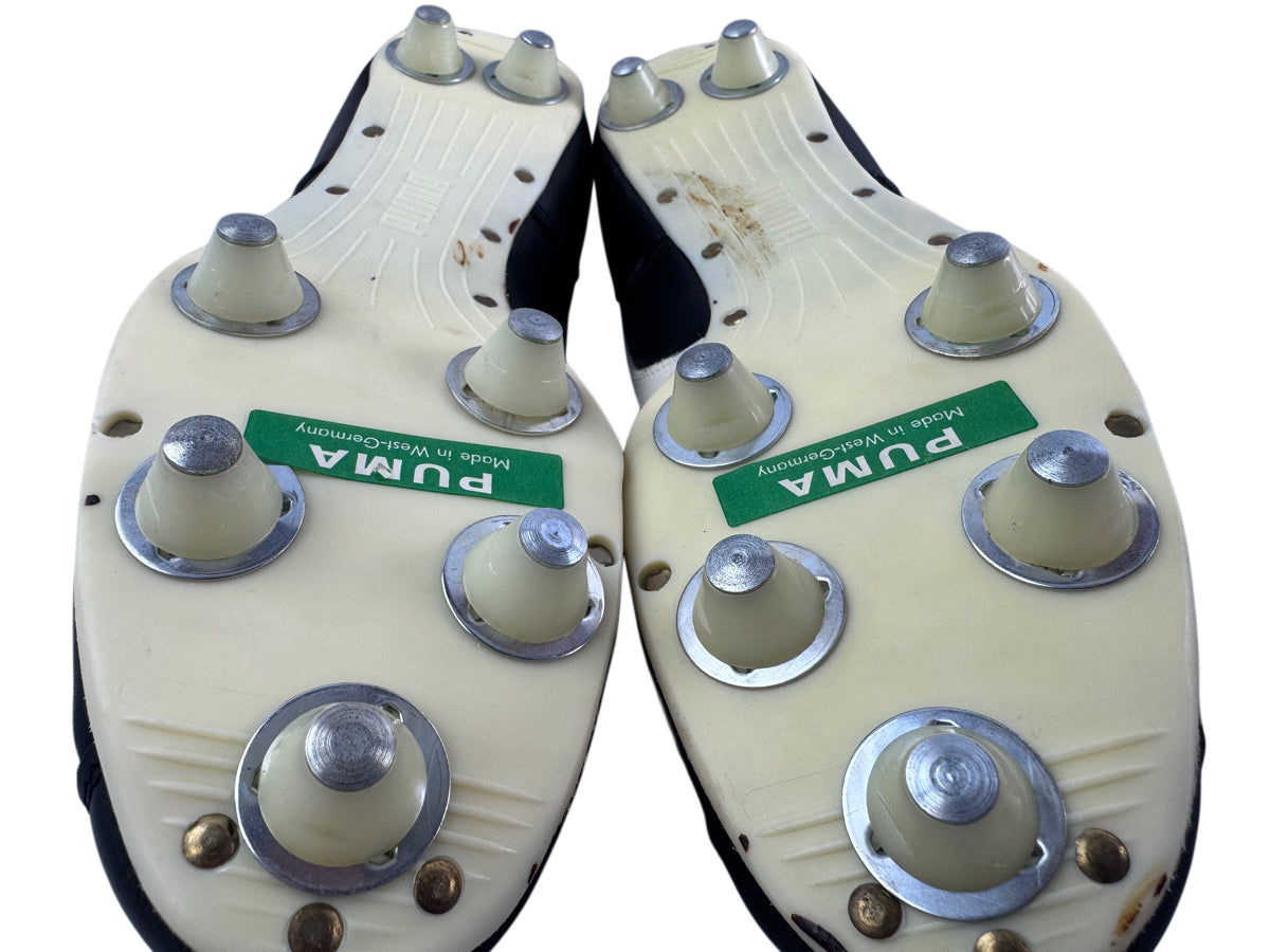 A pair of PUMA soccer/football cleats shown from the bottom, featuring off-white plastic bases with multiple plastic cleats with metal tips and a green PUMA brand label.