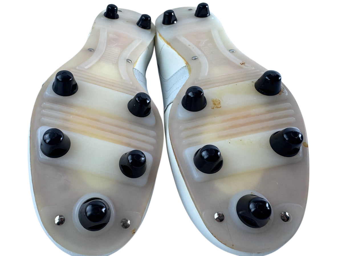 The soles of a pair of white athletic cleats, showing translucent beige and white rubber plates with black rubber studs, photographed on a white background.